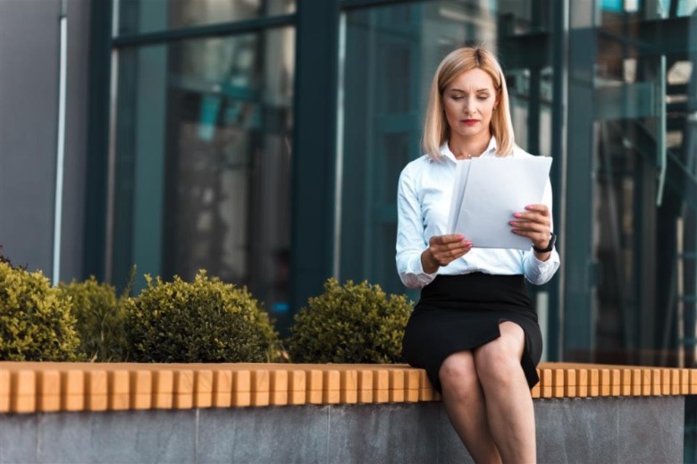 Female office worker sits on a bench near building and looks at documents